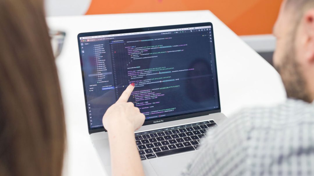 Two people discussing code on a laptop screen, one pointing to the code.
