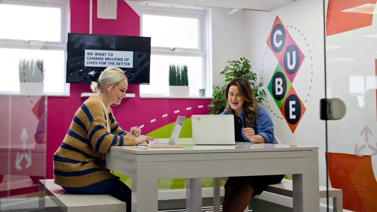 Two women smiling and working on laptops at a table in a colorful office with a motivational screen and wall art in the background.