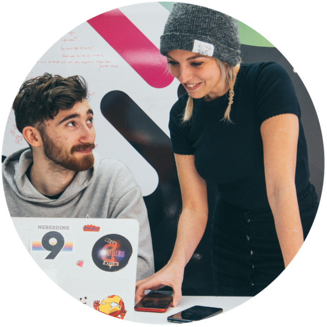 two people collaborating on a project, one sat at a laptop, the other stood holding a mobile device and looking at the laptop screen